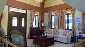 A spacious living room with a sofa, armchair, and coffee table. There is a chandelier, curtains, and a door with glass panels. - Villa Elounda Blue in Plaka (Agios Nikolaos)