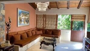 A living room with a brown sectional sofa, a glass coffee table, a chandelier, a painting of a town, and a large window with a view of greenery. - Villa Elounda Blue in Plaka (Agios Nikolaos)
