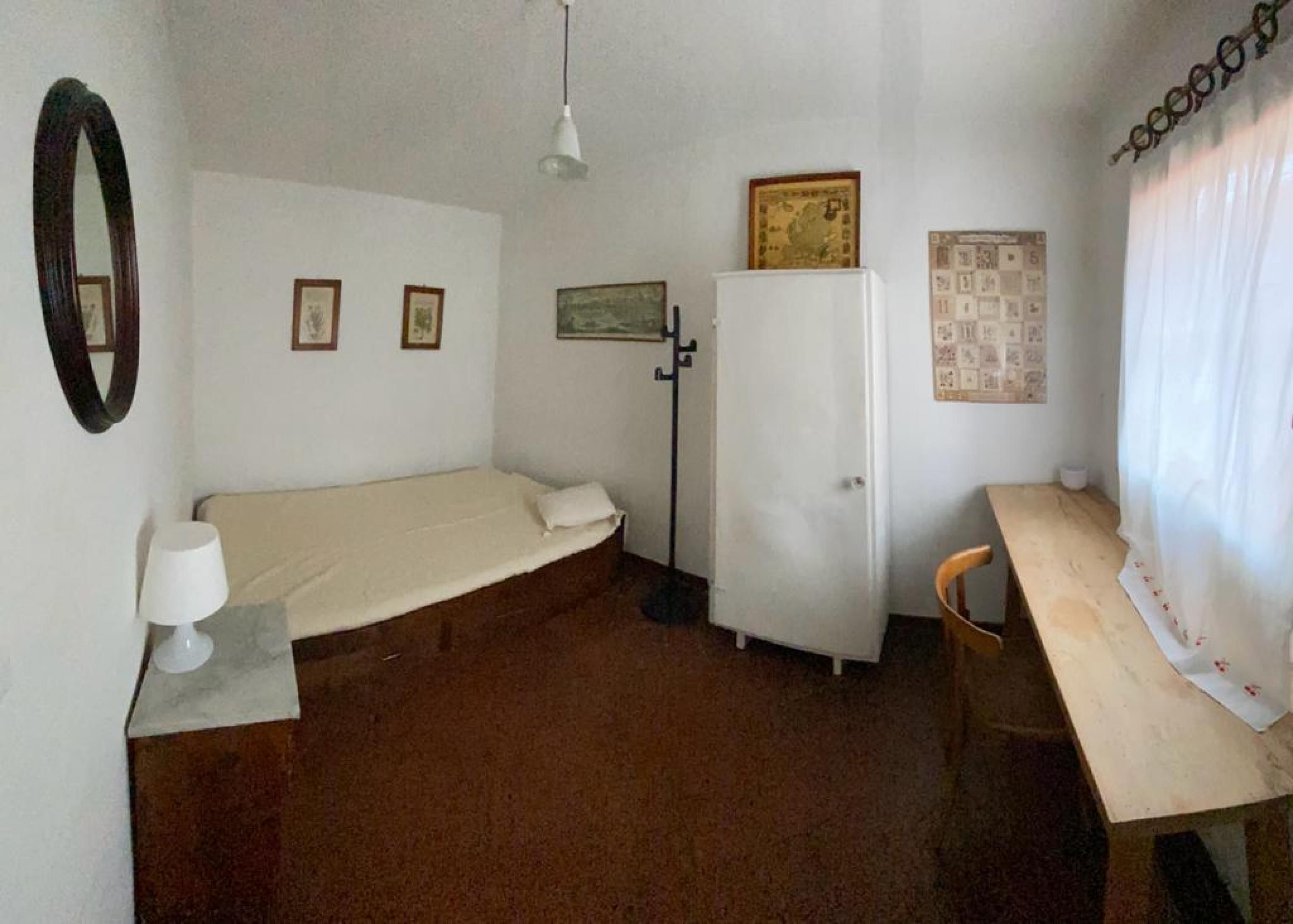 Une petite pièce simple, avec un lit, un bureau, un réfrigérateur, un miroir et quelques tableaux accrochés au mur.