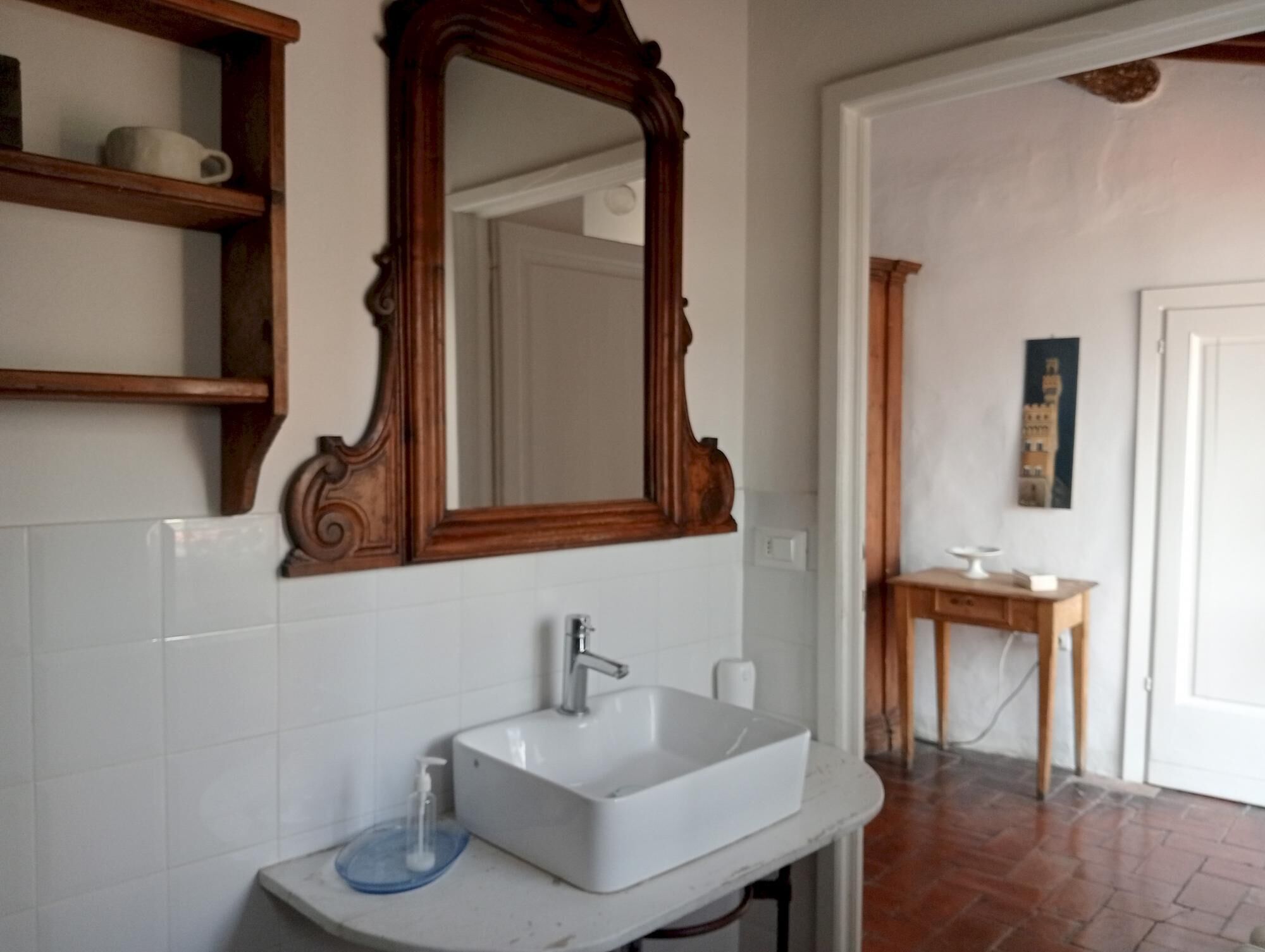 Une salle de bain avec un lavabo blanc, un miroir en bois et une petite table en bois sur laquelle se trouvent une tasse et un livre.