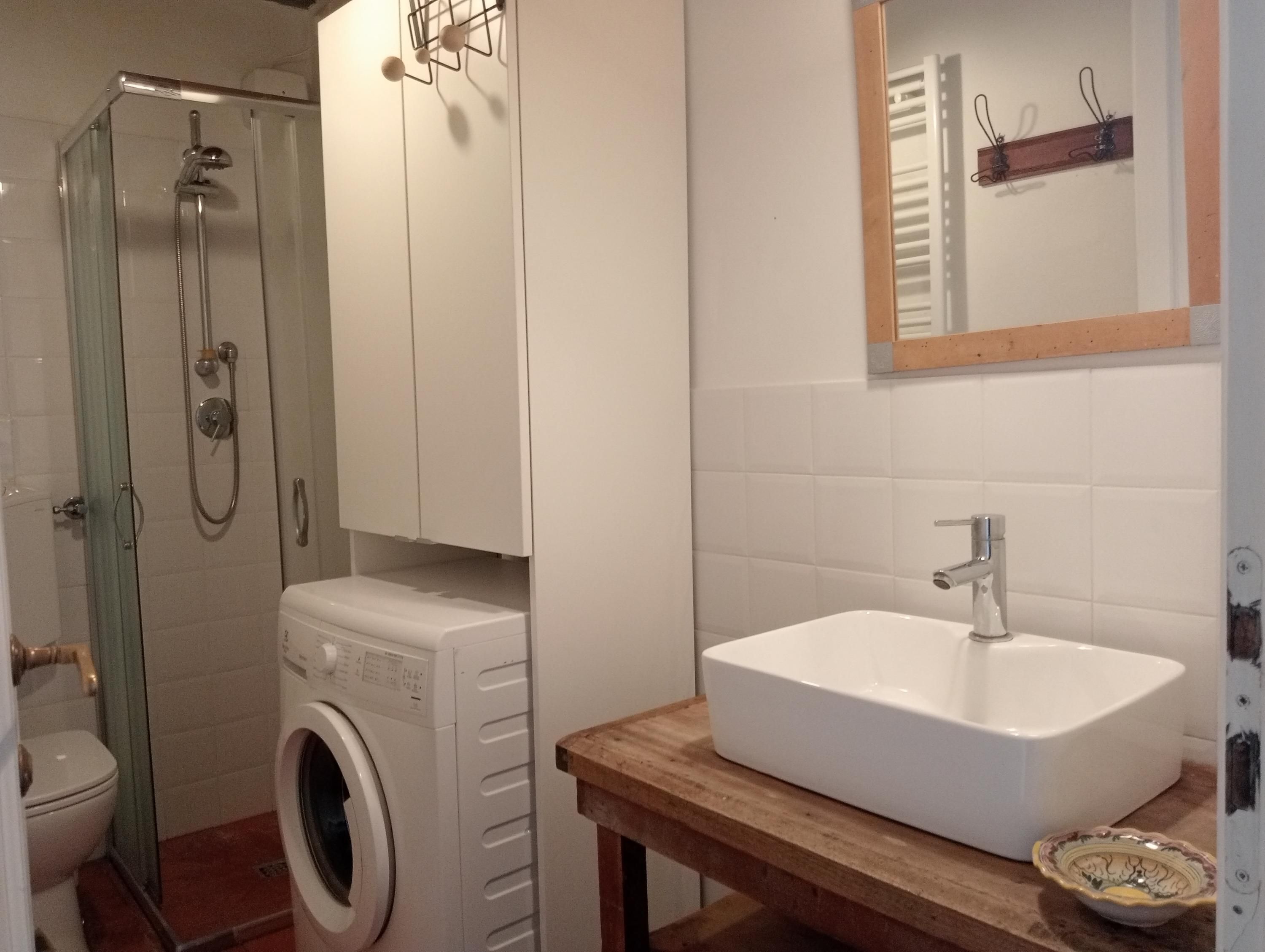 A compact bathroom with a glass-enclosed shower, a built-in washing machine, a wooden vanity with a sink, and a mirror above the vanity.