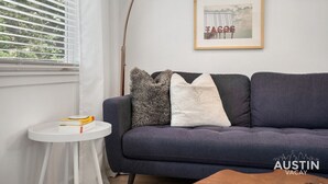 A modern living room with a dark blue sofa, a small white side table with books, a framed picture on the wall, and a window with blinds. - Stylish Austin Retreat w/ Luxurious King Bed + W/D (Austin)