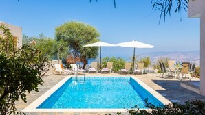 Ein Swimmingpool mit Liegestühlen und Sonnenschirmen, umgeben von viel Grün und freiem Blick auf die Berge.