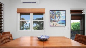Un comedor con mesa de madera, sillas, un cuenco, una ventana con persianas y un cuadro de una playa.