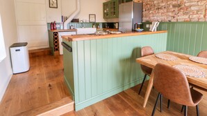 A kitchen with a green island, wooden beams, a brick wall, and a dining area with a table and chairs. - Old Wool Shop (Leyburn)