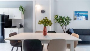 Et moderne køkken med hvide skabe, et sort køleskab og en kaffemaskine. Der er et spisebord med stole og to potteplanter. Et ur hænger på væggen, og et billede er anbragt på væggen til højre.