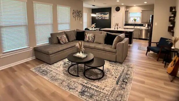 A modern living room with a sectional sofa, a coffee table, and a rug. There is a ceiling fan, a kitchen in the background, and a dining area.