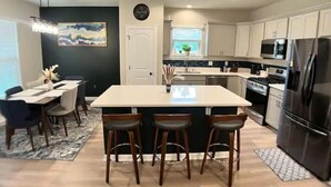 A modern kitchen with a central island, bar stools, a dining area with a table and chairs, and a clock on the wall.