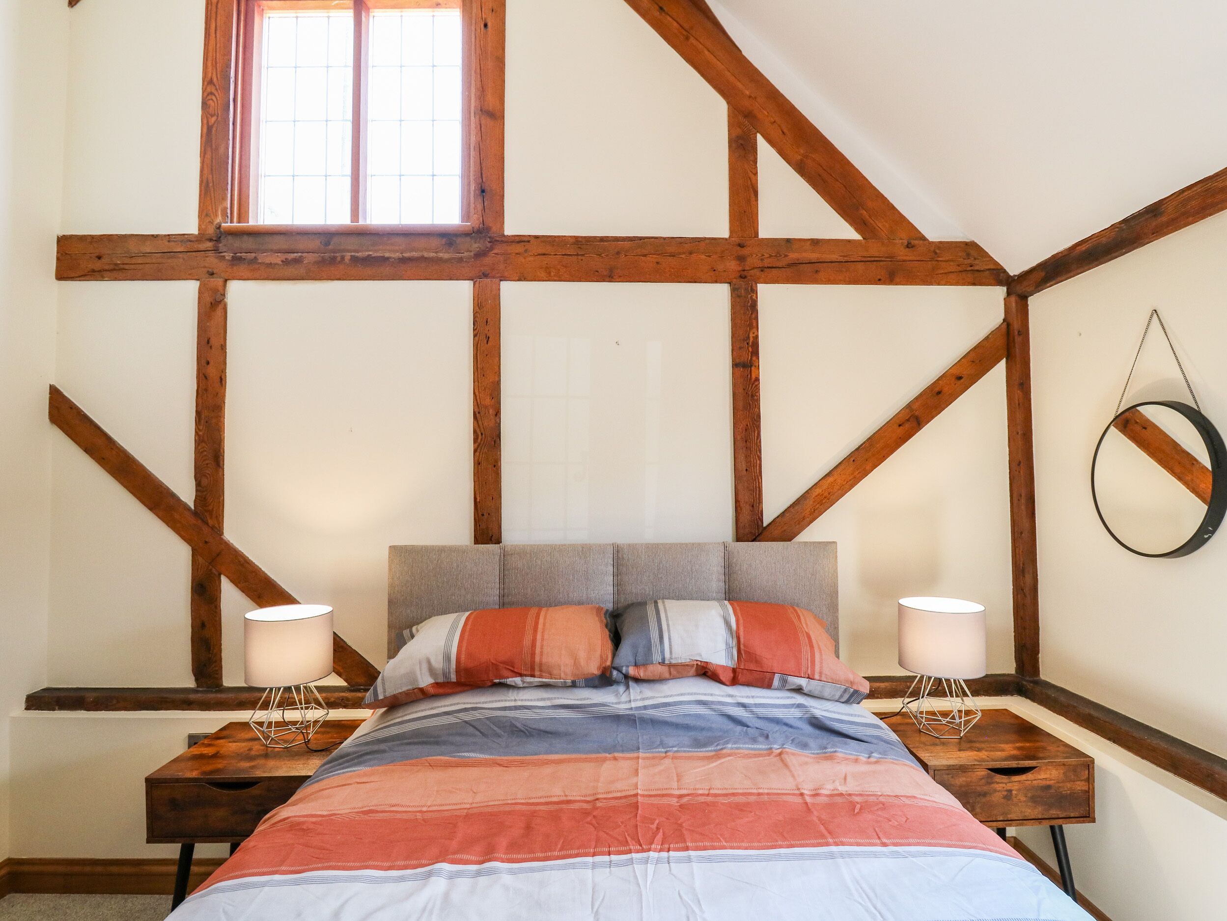 Un dormitorio con vigas de madera a la vista, una cama con ropa de cama a rayas, dos mesitas de noche con lámparas y una ventana con barrotes.