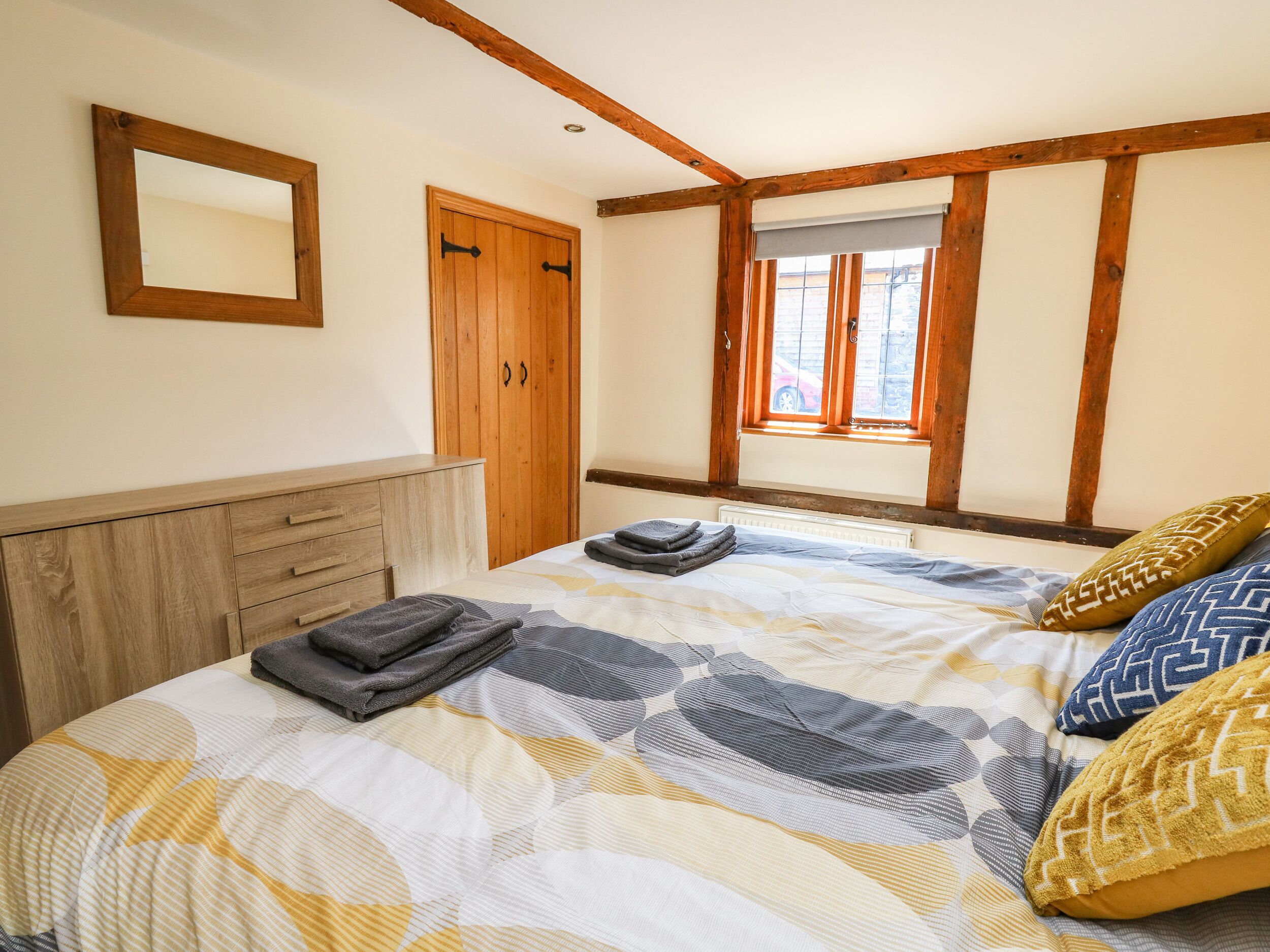 A bedroom with a bed, a wooden wardrobe, a window with blinds, and a mirror on the wall.