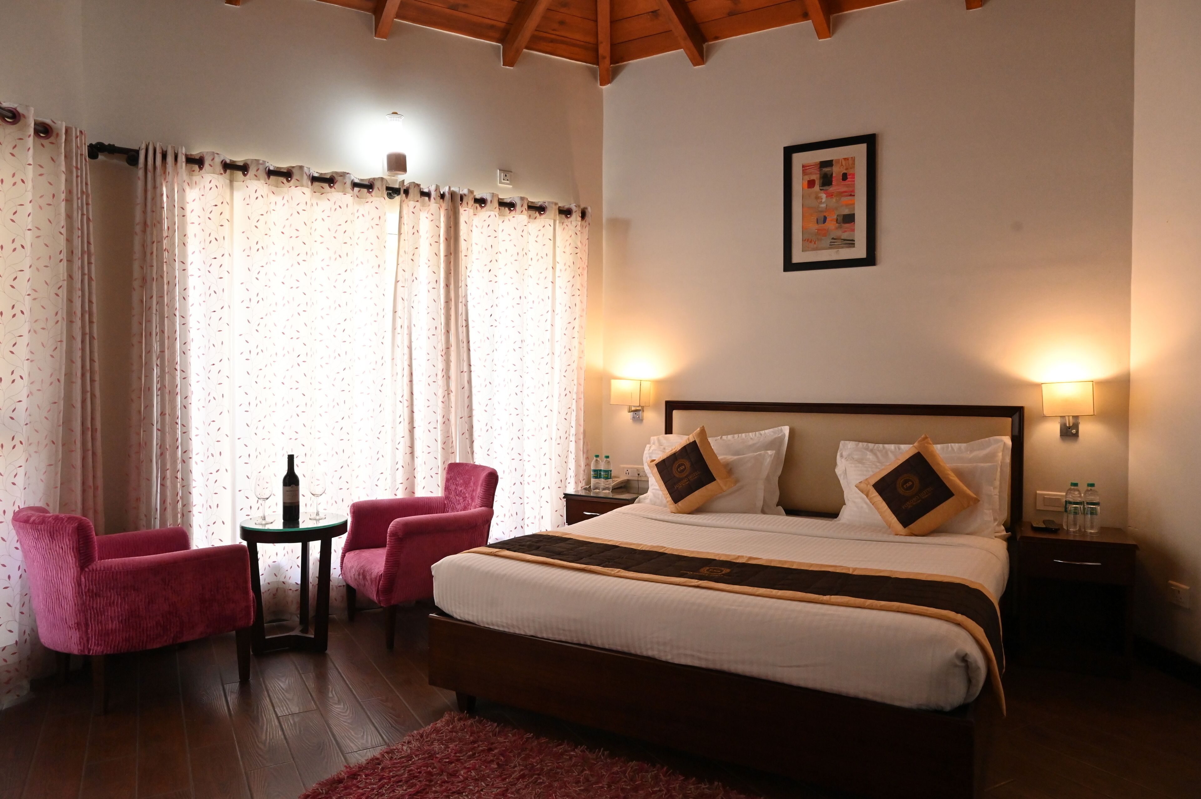 A hotel room with a large bed, two bedside tables, a small table with a bottle and glasses, two pink chairs, and a window with curtains.