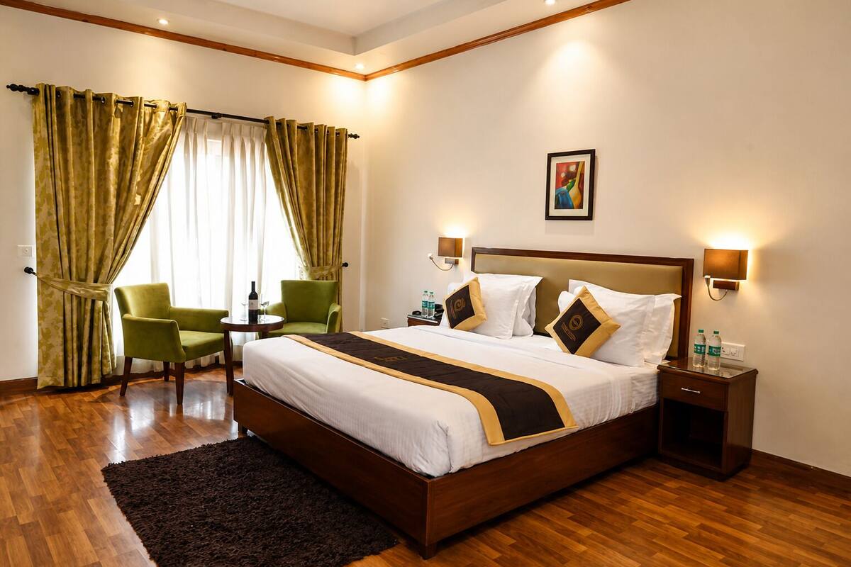 Une chambre d’hôtel avec un grand lit, des fauteuils verts, une petite table avec une bouteille et un tableau au mur.