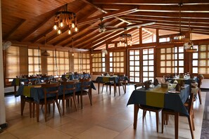 Un vaste espace dédié à la salle à manger, avec des tables et des chaises en bois, de grandes fenêtres et des luminaires suspendus.