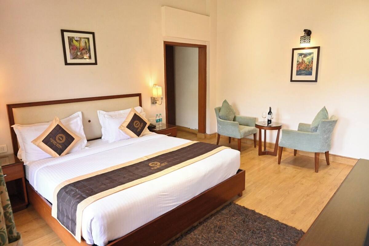 Une chambre d’hôtel avec un grand lit, deux fauteuils, une petite table avec une bouteille et un verre, et deux lampes fixées au mur.