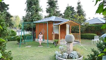 Una pequeña estructura de madera con porche, rodeada por un jardín con diversas plantas y un columpio.