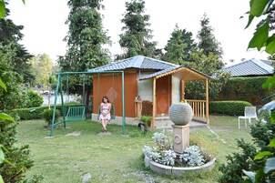 Una pequeña estructura de madera con porche, rodeada por un jardín con diversas plantas y un columpio.