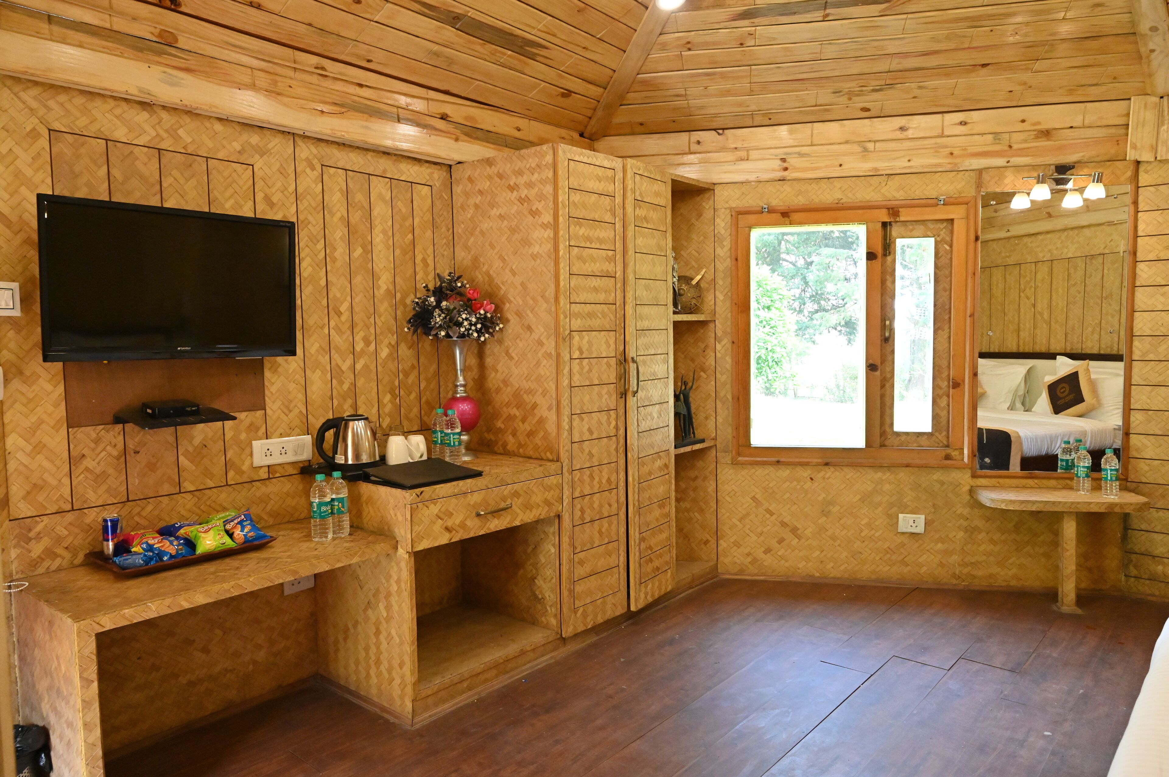 Una cabaña de madera con televisor, un escritorio con aperitivos, un florero con flores y una ventana que ofrece vistas a árboles.