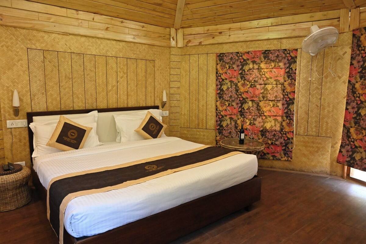 Une pièce aux murs lambrissés, avec un grand lit, un ventilateur fixé au mur et un papier peint à motifs floraux.