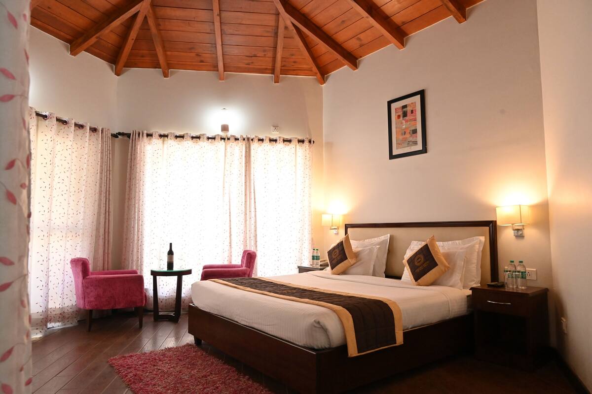 Une chambre d’hôtel avec un plafond en bois, un grand lit avec des oreillers, une table de chevet avec une lampe, une chaise et une petite table sur laquelle se trouve une bouteille.