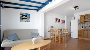 Una sala de estar con sofá, una mesa de comedor de madera y sillas. Se observa una zona de cocina con armarios y un refrigerador.