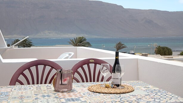 Una mesa con una botella de vino, una copa y una vela sobre un mantel estampado, con vistas a un cuerpo de agua y una montaña.