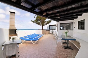 A covered outdoor area with lounge chairs, a table, and a view of the sea.