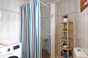 A bathroom with a striped shower curtain, a washing machine, a wooden shelf with toiletries, and a tiled wall.