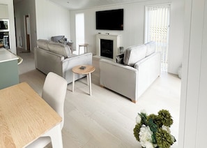 A modern living room with a sofa, a small table, a flat-screen TV mounted above a fireplace, and a dining area with a table and chairs.