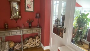 A room with a wooden floor, a red wall, a white door, a mirror, a framed picture, a lamp, a vase with flowers, and a window with a plant outside. - Ground floor Spacious double bed room, free parking close to seafront. (Bexhill)