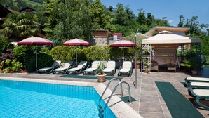 A swimming pool with lounge chairs and umbrellas, surrounded by greenery and buildings. - Holiday Apartment "Sonnegg" with Mountain View, Balcony & Wi-Fi (Saltaus)