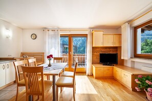 Une cuisine moderne avec des meubles en bois, un coin repas avec des chaises, un téléviseur et une grande fenêtre donnant sur les arbres.