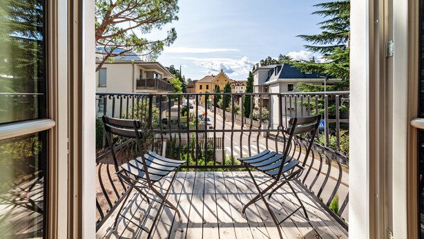 Een balkon met twee stoelen, een leuning en uitzicht op een straat met gebouwen en bomen.