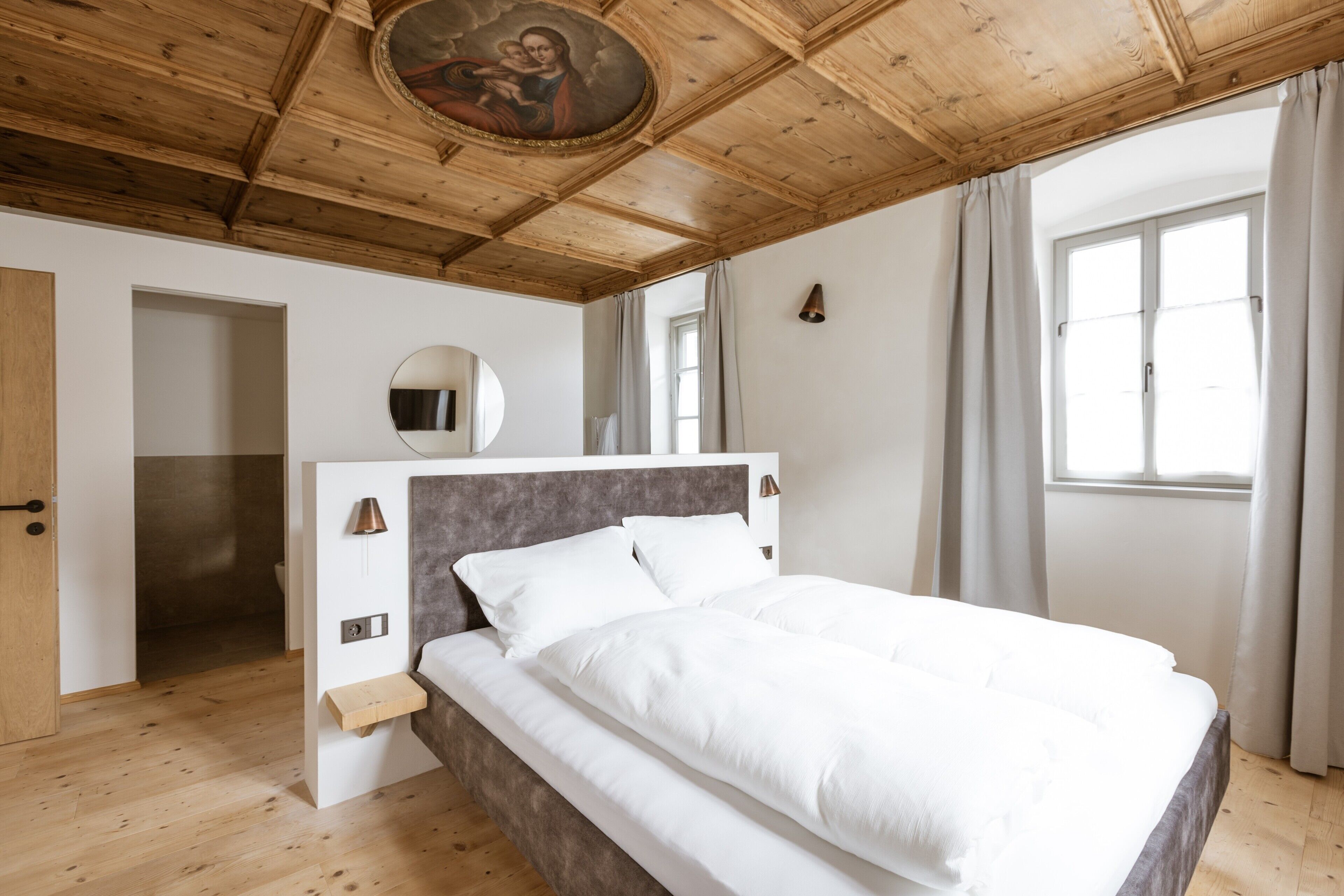 Quarto com teto de madeira, cama com roupa de cama branca, espelho redondo e janela com cortinas.