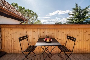 An outdoor wooden deck with a table and two chairs, a bowl of fruit, and a view of trees and sky.