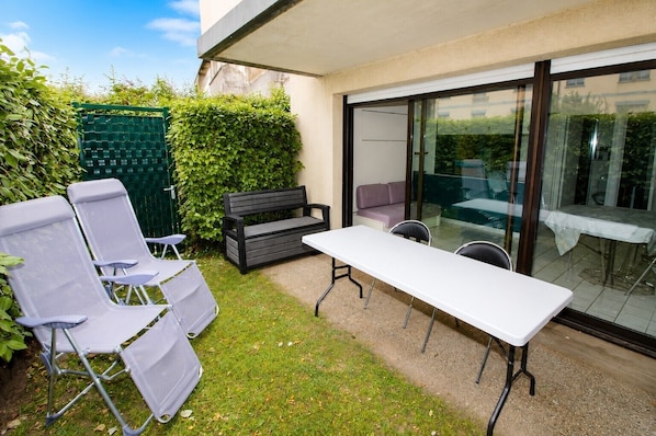 Eine Terrasse im Freien mit einem weißen Tisch, zwei grauen Lounge-Stühlen, einem schwarzen Sofa und einer Glastür, die zu einem überdachten Bereich führt.