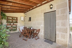 Eine überdachte Terrasse mit Steinboden, Holzbalken an der Decke, einem Tisch mit Stühlen, Topfpflanzen und einer Wandleuchte.