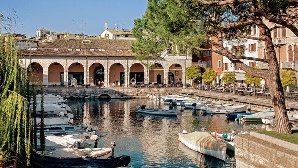 A waterfront area with boats docked, a stone building with arches, and a tree. - Apartment 'Casa Cattaleya' with Shared Pool, Private Terrace and Shared Garden (Desenzano del Garda)