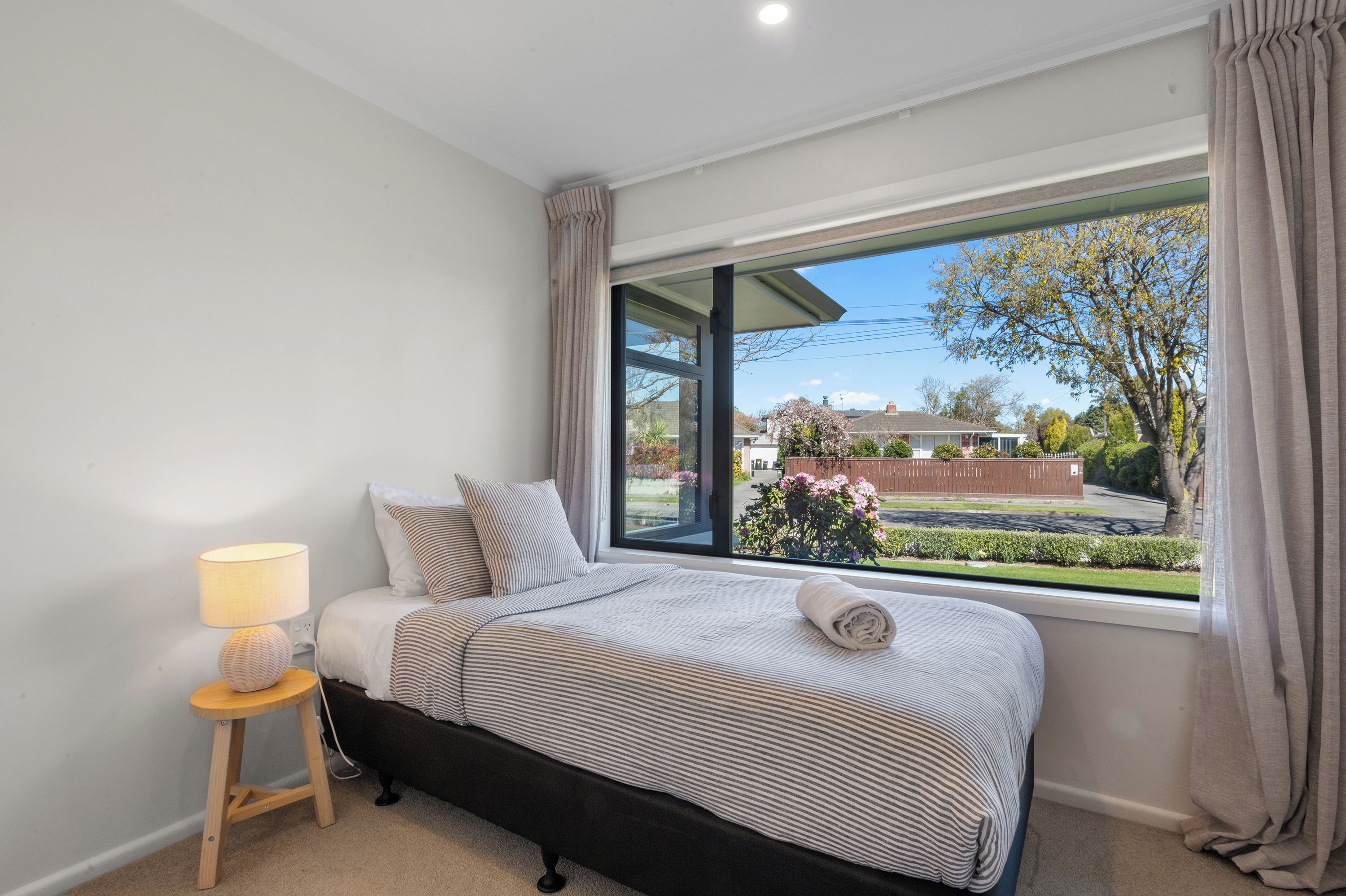 A bedroom with a large window, a bed with striped bedding, a bedside lamp, and a view of a garden and neighboring houses.