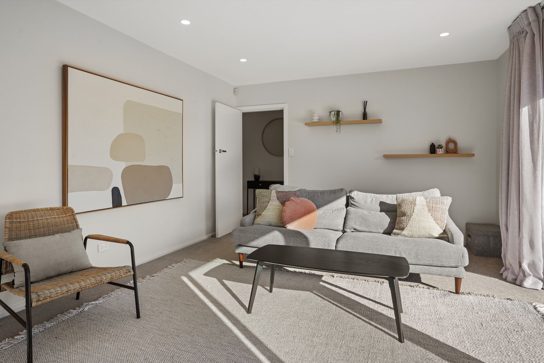 A modern living room with a grey sofa, a wicker chair, and a black coffee table. There are two wooden shelves on the wall, and a large abstract painting above the sofa.