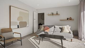 A modern living room with a grey sofa, a wicker chair, and a black coffee table. There are two wooden shelves on the wall, and a large abstract painting above the sofa.