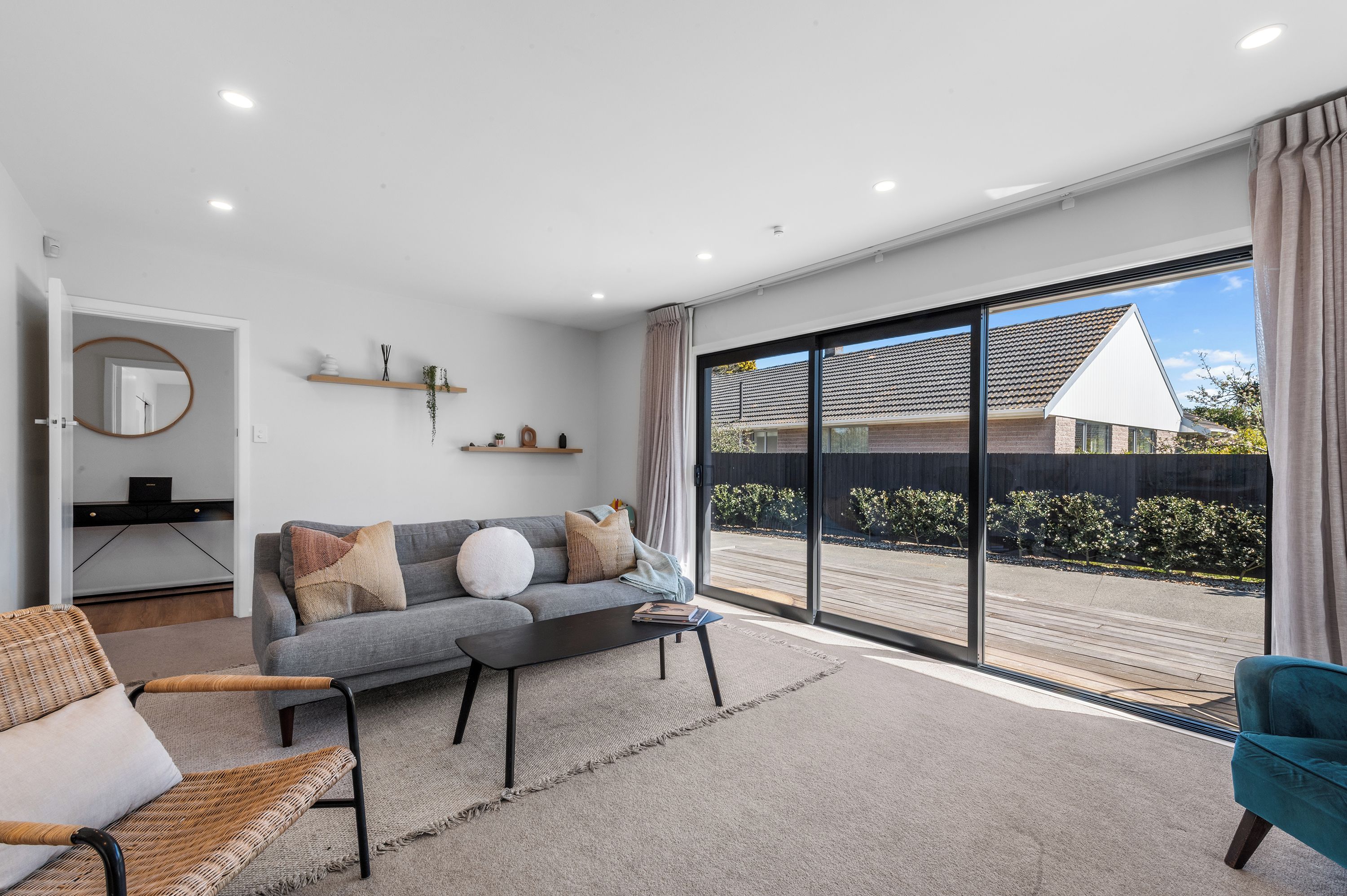 A modern living room with a grey sofa, a coffee table, and a wicker chair. There is a large sliding glass door leading to a balcony with a view of a house and trees.