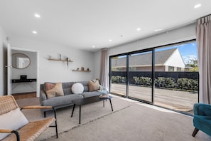 A modern living room with a grey sofa, a coffee table, and a wicker chair. There is a large sliding glass door leading to a balcony with a view of a house and trees.