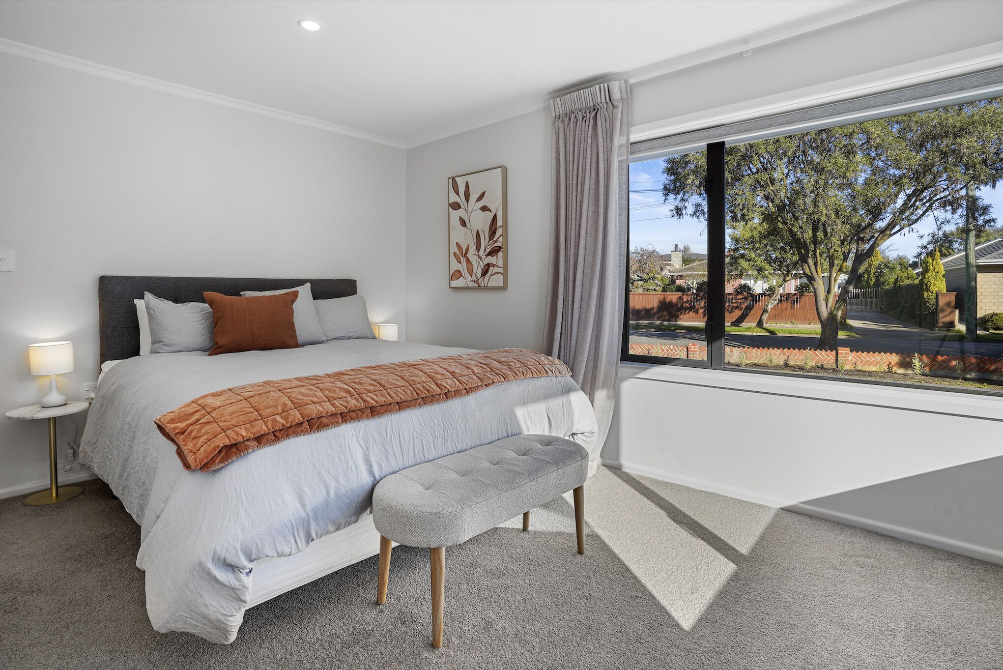 A bedroom with a large bed, a bench, a nightstand, a lamp, and a window with a view of a tree and a fence.