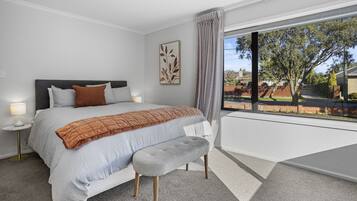 A bedroom with a large bed, a bench, a nightstand, a lamp, and a window with a view of a tree and a fence.