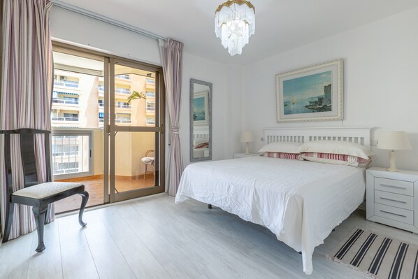 Une chambre avec un grand lit, une chaise, une table de chevet, une lampe, un tableau et une vue sur l’extérieur.