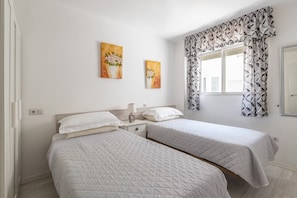 A bedroom with two beds, a window with patterned curtains, and two wall-mounted paintings.
