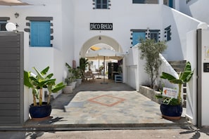 A hotel entrance with a sign reading 'Pico Bello,' a paved pathway, potted plants, and a seating area.