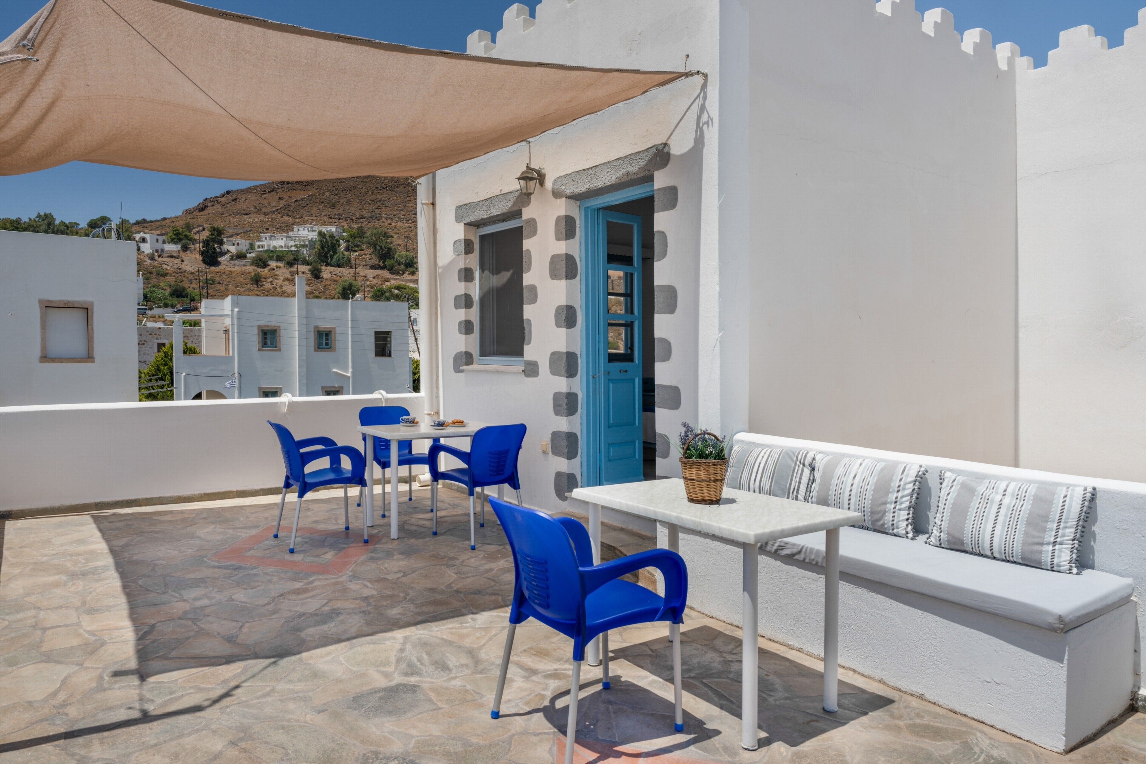 Een terras met een witte tafel, blauwe stoelen, een witte bank en een beige zonnescherm.
