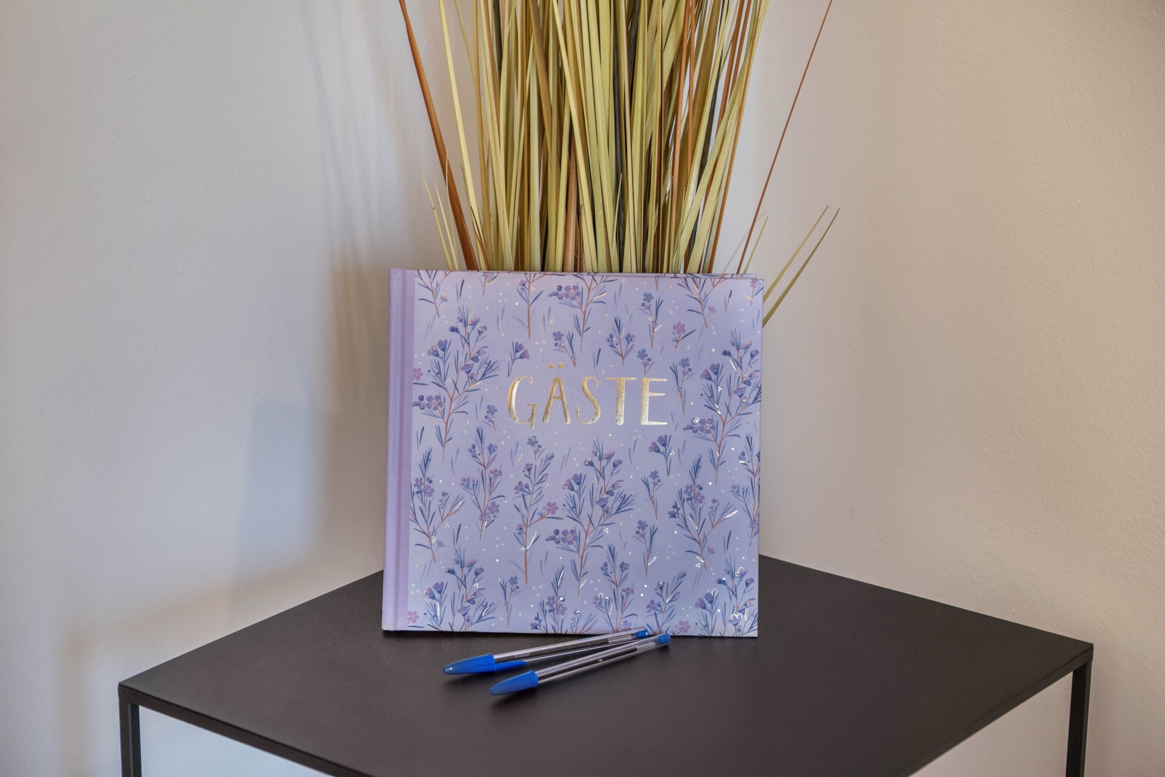 Ein Buch mit dem Titel 'GASTE' auf einem schwarzen Tisch, daneben liegen blaue Stifte.