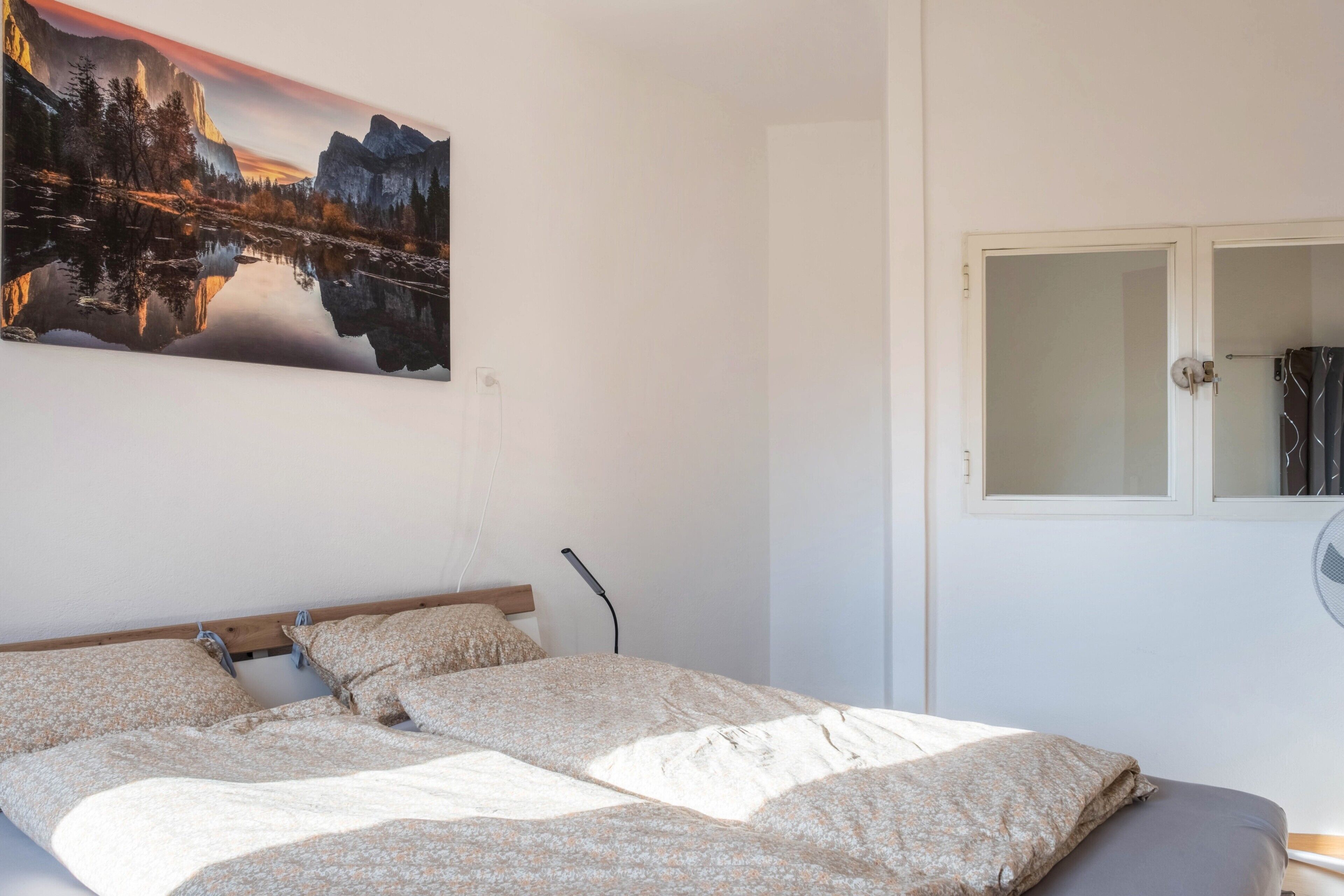 Une chambre à coucher avec un lit, un tableau représentant un paysage de montagne, une fenêtre donnant sur un beau panorama et une table de chevet.